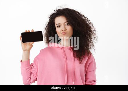 Disappointed curly brunette girl sulking, showing horizontal smartphone screen with upset face, complaining on something online, standing against Stock Photo