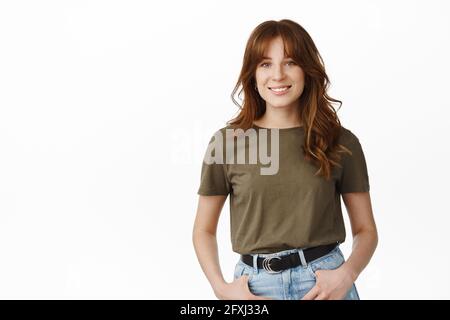 Portrait of smiling redhead girl with bangs, looking cute and happy at camera, casually standing, relaxed pose with hands in jeans pockets, white Stock Photo