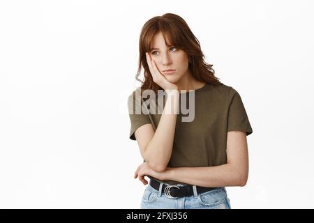 Image of bored and gloomy young woman, student lean face on hand and look uninterested aside, attend boring meeting, feeling sad, standing against Stock Photo