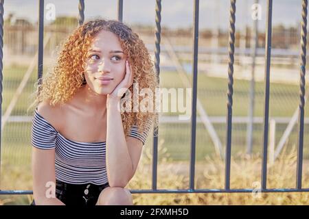 Young curly haired latina thought sitting on a fence. Cute latina girl in black shorts and striped t-shirt. High quality photo Stock Photo