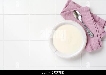 Greek yogurt in white bowl, linen napkin and wooden spoon on white table. Top view Stock Photo