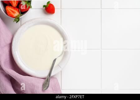 Greek yogurt in white bowl with ingredients for making breakfast granola and fresh strawberries on white table. Top view Stock Photo