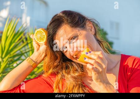 Beautiful woman portrait eating a lemon slice.Female biting a lemon in red shirt outdoor in nature background.. Stock Photo