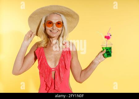 Cheerful blonde woman in sunglasses, red swimsuit and straw hat smiling at camera, adjusting her hat and holding cocktail drink while posing isolated Stock Photo