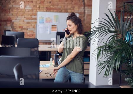 Young woman, architect or interior designer looking away while talking on the phone, standing in modern green office and discussing new project Stock Photo