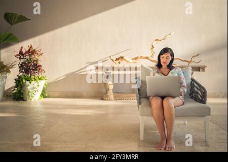 Female freelance worker sitting in a cozy armchair is using a laptop Stock Photo