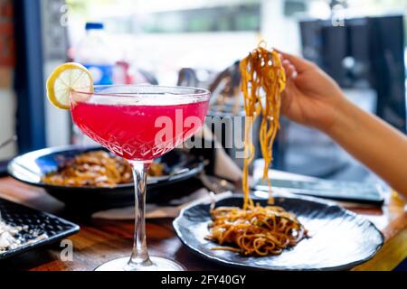 Wide stemmed wine glass filled with red pink strawberry cherry bloody mary drink with slice of lime with out of focus woman in background picking up Stock Photo