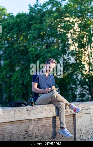 Rome, Italy - May 26, 2021: A middle-aged man sitting on the low wall in the streets of the historic center, writes notes in his diary. Stock Photo