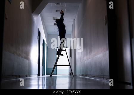 Technician Repairing Ceiling Fixture In Office On Stepladder Stock Photo