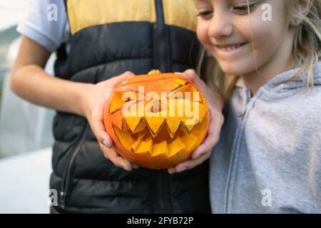 Child holding and open top of a carved pumpkin with ghoulish face, jack-o-lantern, in the background boy sitting at the table and decorating pumpkin Stock Photo