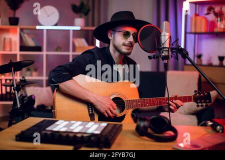 Handsome man singing in microphone and playing guitar while recording song at modern studio. Young singer in trendy hat and sunglasses creating new track indoors. Stock Photo