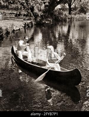 1930s COUPLE MAN AND WOMAN PADDLING A CANOE GOING ON A SUMMER PICNIC WITH BASKET AND THERMOS  - c1605 HAR001 HARS FITNESS ROWING BLOND HEALTHY YOUNG ADULT TEAMWORK CANOE PLEASED JOY LIFESTYLE FEMALES MARRIED RURAL SPOUSE HUSBANDS COPY SPACE FRIENDSHIP HALF-LENGTH LADIES PERSONS STREAM MALES TRANSPORTATION B&W PARTNER SUMMERTIME CANOEING ACTIVITY PHYSICAL CHEERFUL HIGH ANGLE ADVENTURE STRENGTH AND EXCITEMENT RECREATION THERMOS OARS SMILES AL FRESCO PADDLES FLEXIBILITY JOYFUL MUSCLES PICNICKING STYLISH COOPERATION PADDLING SEASON TOGETHERNESS WIVES YOUNG ADULT MAN BLACK AND WHITE Stock Photo