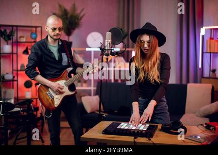 Handsome man playing electro guitar while woman recording and mixing sound on dj controller. Group of professional musician making modern music at studio. Stock Photo