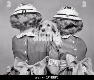 1950s TWO TWIN GIRLS BACK TO CAMERA FANCY HATS AND DRESSES HOLDING COCKER SPANIEL PUPPY LOOKING AT CAMERA BETWEEN THEM  - d2628 HAR001 HARS MATCH FANCY PETS COCKER B&W DRESSES MATCHING SAME MAMMALS AND CANINES REAR VIEW TO BETWEEN SIBLING POOCH THEM FROM BEHIND BOWS LOOK-ALIKE BACK VIEW CANINE DUPLICATE JUVENILES LOOK ALIKE MAMMAL PUP BLACK AND WHITE CAUCASIAN ETHNICITY HAR001 OLD FASHIONED Stock Photo