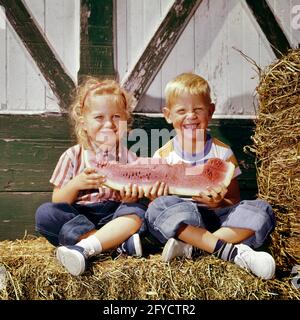 1960s BLONDE BOY AND GIRL BROTHER SISTER SITTING ON A HAY BALE LOOKING AT THE CAMERA SMILING SHARING A LARGE SLICE OF WATERMELON - kf3085 HAR001 HARS BROTHER OLD FASHION SISTER 1 JUVENILE CUTE STYLE BLOND FRIEND HAY TEAMWORK COTTON PLEASED JOY LIFESTYLE SATISFACTION FEMALES BROTHERS BARN RURAL HEALTHINESS HOME LIFE COPY SPACE FRIENDSHIP HALF-LENGTH FARMING MALES SNEAKERS SIBLINGS DENIM SISTERS AGRICULTURE EYE CONTACT SLICE HAPPINESS CHEERFUL AND FARMERS NUTRITION SIBLING SMILES WATERMELON CONNECTION CONSUME CONSUMING FRIENDLY JOYFUL NOURISHMENT BALE BLUE JEANS COOPERATION GROWTH INFORMAL Stock Photo