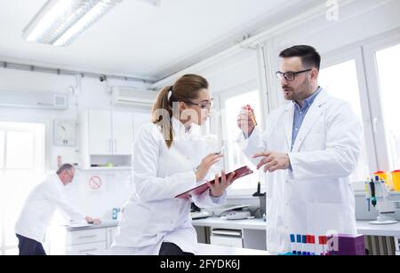 Two young lab technicians scientists talking experimenting in laboratory. Man and woman doctors holding vial clipboard doing research. Stock Photo