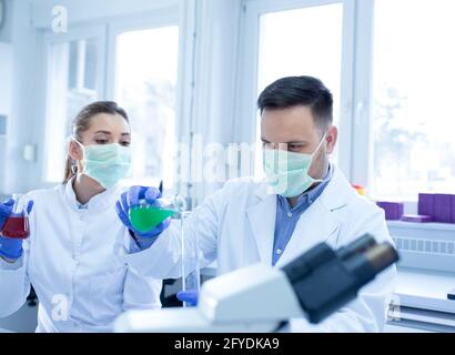 Young scientists researching experimenting holding erlenmeyer flask. Man and woman lab technician in laboratory measuring liquid wearing face mask. Stock Photo