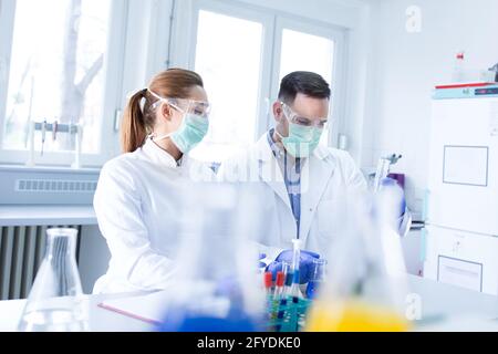 Young scientists researching experimenting using vial and beaker. Man and woman lab technician looking monitoring wearing face mask and safety glasses Stock Photo