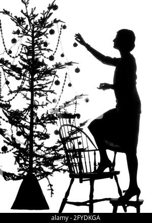 1920s SILHOUETTE OF ANONYMOUS YOUNG WOMAN STANDING ON CHAIR HANGING FESTIVE FANCY BALL DECORATIONS ON TALL CHRISTMAS PINE TREE  - s1468 HAR001 HARS CELEBRATION FEMALES STOOL HOME LIFE COPY SPACE FULL-LENGTH LADIES PERSONS INSPIRATION CARING DANGER FANCY SPIRITUALITY CONFIDENCE B&W HAPPINESS STRENGTH SILHOUETTED MERRY CHOICE LOW ANGLE RECREATION PRIDE OF ON DECEMBER UNSAFE CONCEPTUAL DECEMBER 25 STYLISH ANONYMOUS WINDSOR JOYOUS MID-ADULT MID-ADULT WOMAN YOUNG ADULT WOMAN BLACK AND WHITE FESTIVE HAR001 OLD FASHIONED Stock Photo