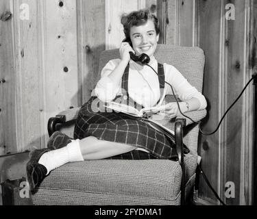 1950s TEEN GIRL SMILING TALKING ON THE TELEPHONE READING BOOK IN HER LAP WEARING BOBBY SOCKS LOAFERS SITTING IN EASY CHAIR - t2012 HAR001 HARS PLEASED JOY LIFESTYLE SATISFACTION HEALTHINESS HOME LIFE COMMUNICATING COPY SPACE FRIENDSHIP HALF-LENGTH PERSONS TEENAGE GIRL B&W LAP FREEDOM SCHOOLS BUZZ HAPPINESS CHEERFUL EXCITEMENT BUSYBODY TELLING HIGH SCHOOL PHONES SMILES STORIES HIGH SCHOOLS CONNECTION CONCEPTUAL LOAFERS TELEPHONES BOBBY EASY JOYFUL STYLISH TEENAGED BOBBY SOX KNOTTY PINE TATTLER HEARSAY PANELING RUMORS BLACK AND WHITE BOBBY SOCKS CAUCASIAN ETHNICITY HAR001 OLD FASHIONED Stock Photo