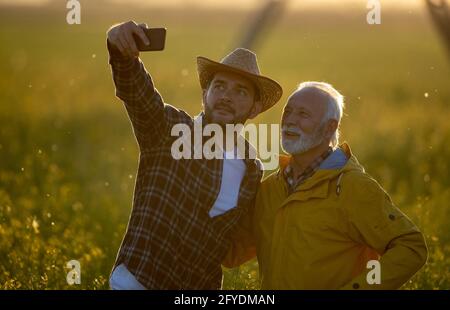 Two farmers standing in field taking selfie using phone smiling happy. Men marking in agribusiness with photo outdoors at sundown. Stock Photo