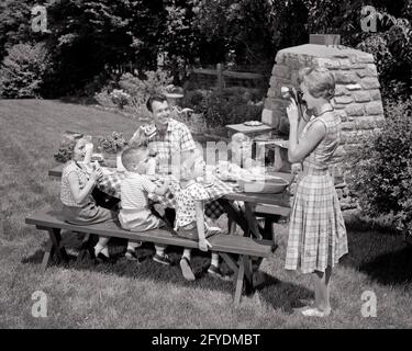 1960s MOTHER TAKING SNAPSHOT PHOTO OF FAMILY PICNIC BACKYARD BBQ KIDS AND DAD AT TABLE  - u25 HAR001 HARS DAD FOUR MOM NOSTALGIC BARBEQUE PAIR 4 COMMUNITY SUBURBAN BACKYARD MOTHERS OLD TIME NOSTALGIA BROTHER BARBECUE OLD FASHION SISTER 1 PHOTOGRAPH JUVENILE TEAMWORK SONS FAMILIES JOY LIFESTYLE SATISFACTION FEMALES MARRIED BROTHERS RURAL SPOUSE HUSBANDS HEALTHINESS HOME LIFE 6 COPY SPACE FULL-LENGTH HALF-LENGTH LADIES DAUGHTERS PERSONS SNAPSHOT MALES SIX SIBLINGS SISTERS FATHERS B&W PARTNER SUMMERTIME HAPPINESS HIGH ANGLE LEISURE AND DADS OPPORTUNITY SIBLING PICNIC TABLE STYLISH BAR-B-QUE Stock Photo