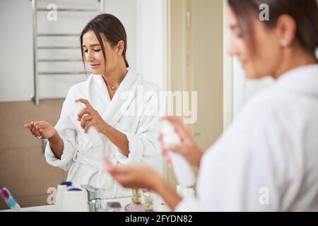 Happy positive young lady using spray on hands while standing in room indoors Stock Photo