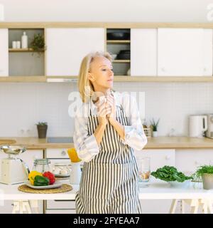 Starting new day. Side view of beautiful thoughtful senior woman drinking coffee or tea and thinking about something, elderly lady in apron enjoying morning while standing in modern kitchen at home Stock Photo