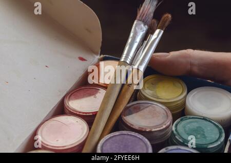 Set of paints for painting in cylindrical plastic containers and brushes. A man's hand holds a box of colorful gouache paints. Tinted photo Stock Photo