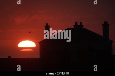 The sun rises through the clouds behind a house on St Mary's Island in Whitley bay on the North East coast. Picture date: Friday May 28, 2021. Stock Photo