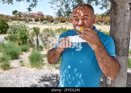 Funny bearded middle aged man eating vegan sandwich on the street under shadow of the tree. Unhealthy eating. Fast food. Takeaway food Stock Photo