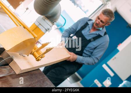 Portrait of an elderly carpenter or carpenter in overalls working with wooden boards in a carpenter's workshop. Sawing wood. Stock Photo