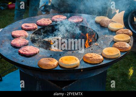 Beef patties and burger buns are grilled on a large, round wood-burning grill outside. Barbecue festival in the city park. Street fast food. Stock Photo