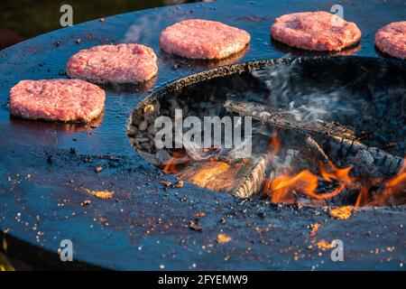 Beef patties for burgers are being grilled on a large round wood-fired grill. Close-up. Barbecue festival in the city park. Street fast food. Stock Photo