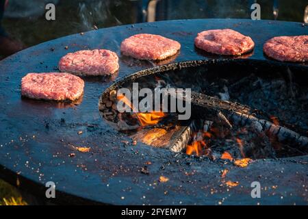 Beef patties for burgers are being grilled on a large round wood-fired grill. Close-up. Barbecue festival in the city park. Street fast food. Stock Photo