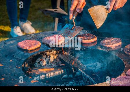 On the lawn, a large round wood-burning grill is roasting beef patties for burgers. The cook turns over the fried cutlets. Barbecue festival in the ci Stock Photo