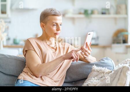 Chatting with friend. Happy attractive mature woman using smartphone while sitting on sofa and enjoying leisure time at home. Selective focus on senior female. Elderly people and modern technologies Stock Photo