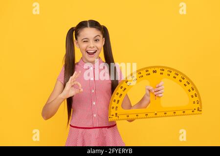 winking teen girl hold school math tool protractor ruler on yellow background show ok, education Stock Photo