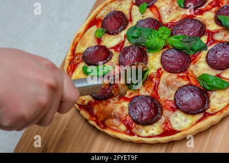 Slicing freshly cooked homemade pizza on a kitchen board topped with cheese, sausage, basil. Stock Photo