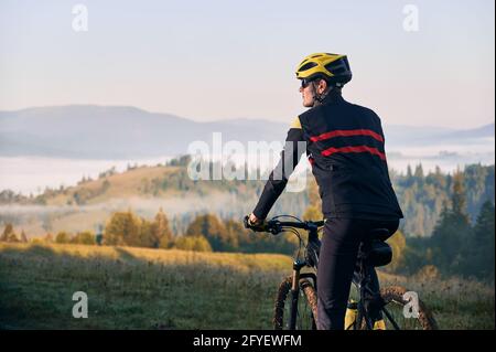 Close up male cyclist in cycling suit riding bike with hills on background. Man bicyclist wearing safety helmet and glasses while enjoying bicycle ride in mountains in the morning. Back view Stock Photo