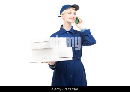 Horizontal medium portrait of modern beautiful Caucasian postal worker wearing blue uniform holding boxes talking on phone, white background Stock Photo