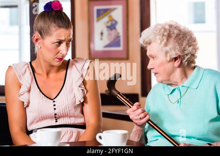 Grandmother and granddaughter having argument in cafe, the senior woman in threatening with her crutch Stock Photo
