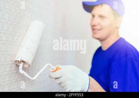 A painter in a cap and gloves paints the wall with a paint roller. Portrait of a young worker in uniform. Professional repair of premises. Mockup Stock Photo