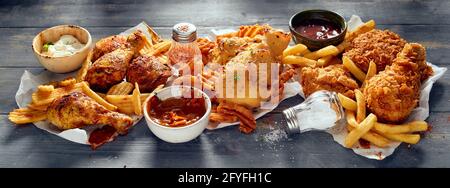 High angle appetizing crispy chicken legs with potato wedges and fries served on tissues near various sauces on black wooden table Stock Photo