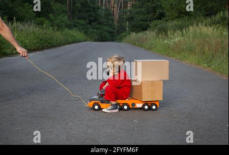 toddler boy in red overalls sits on a big toy car - a truck with cardboard boxes, talking on phone. Parcel delivery, little postman. like Dad. a man's Stock Photo