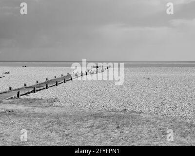 Monochrome image of wooden pathway leading across stony beach to sea Stock Photo