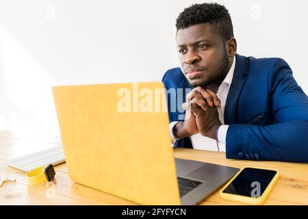 Pensive Afro American male professional freelancer having an online meeting .Concentrated businessman sitting at the desk, using yellow laptop and Stock Photo