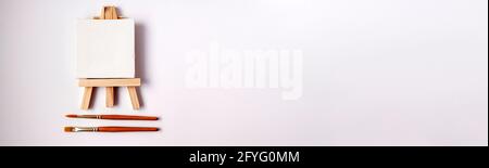 Flat lay banner of artistic orange metal paint brushes and tiny wooden easel and white canvas close up on white background with copy space from above. Stock Photo
