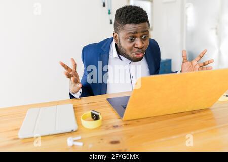 Shocked man is staring on the yellow laptop screen, annoyed African-American businessman received bad news, freelancer guy suffering business problems Stock Photo