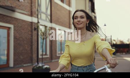 joyful young woman in yellow blouse smiling while riding on bicycle outside Stock Photo